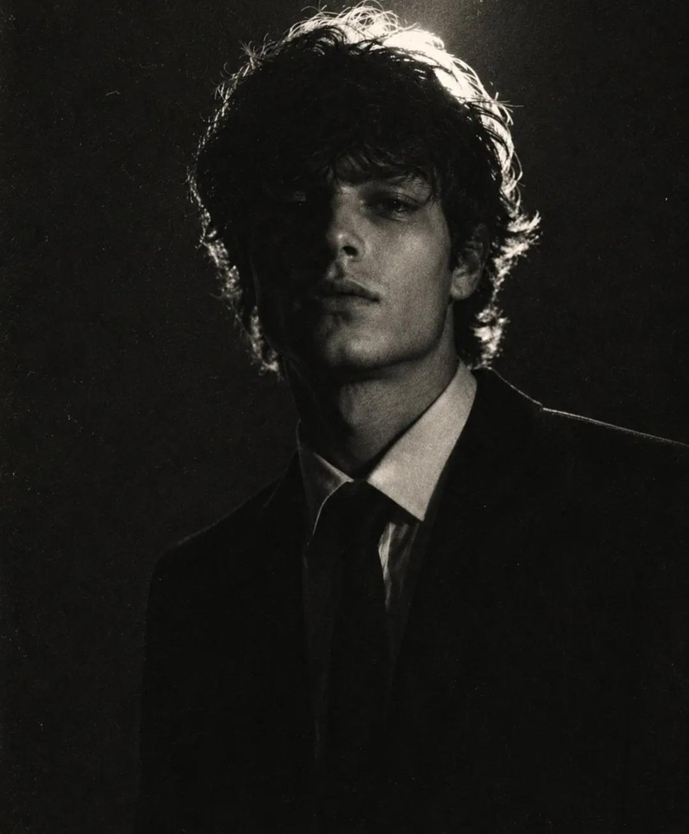 A young man in a black suit stands solemnly in dark studio light.