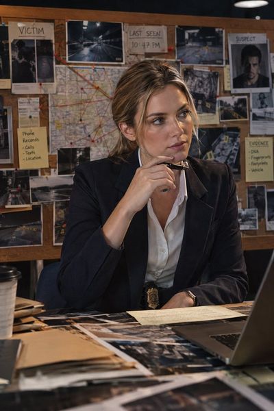 Female detective in navy blazer studies evidence board with serious focus