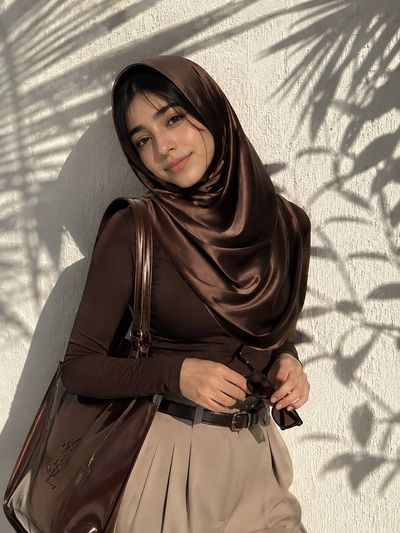 Elegant woman in chocolate hijab leans against sunlit wall