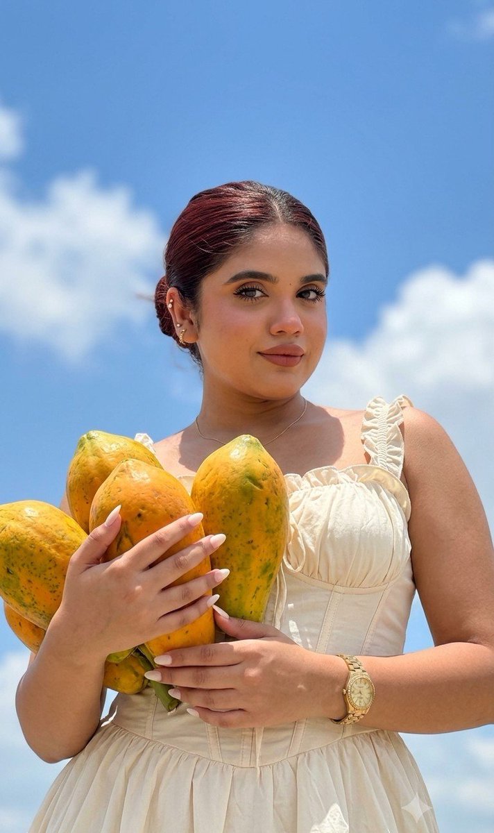 Woman holds papayas under bright blue sky with fluffy clouds.