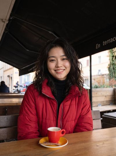 Young East Asian woman smiles brightly in red puffer jacket at cafe