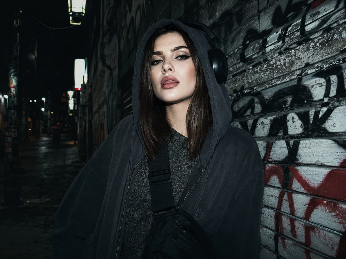 A young woman in an alley poses with headphones and a hoodie