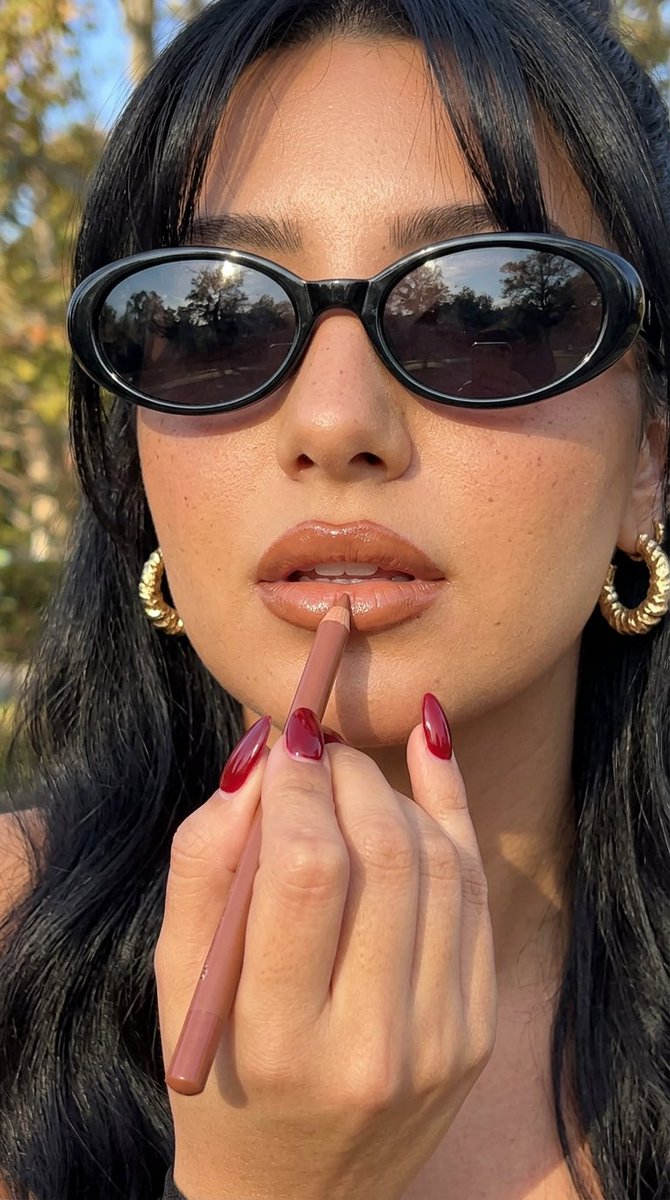 Woman applies caramel lip liner with gold hoop earrings in sunlight
