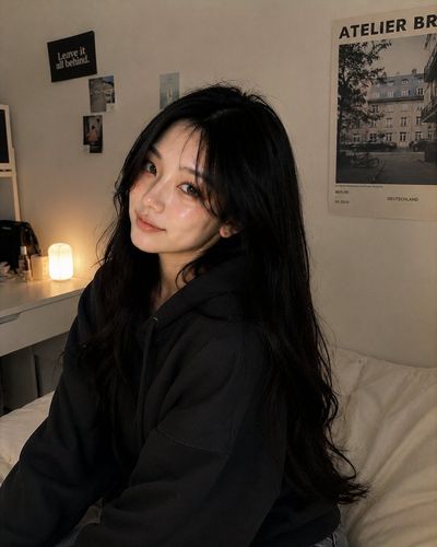Korean girl rests on cream bed in cozy hoodie under warm light