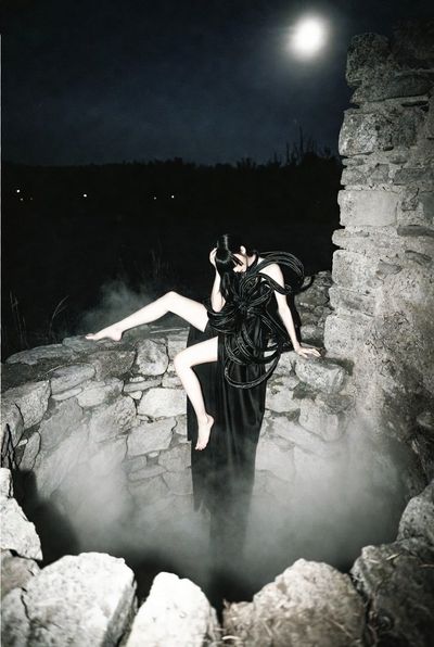 A lone woman in a black dress sits by an ancient well under moonlight