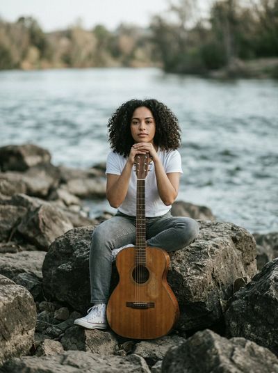 Woman with curly hair holds guitar on rocky riverbank