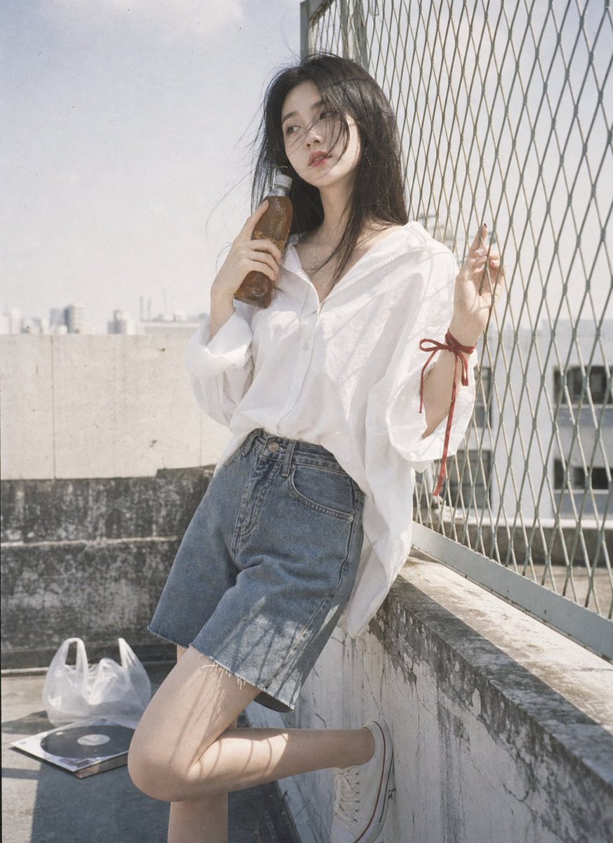 A woman in white leans by a fence with a glass bottle