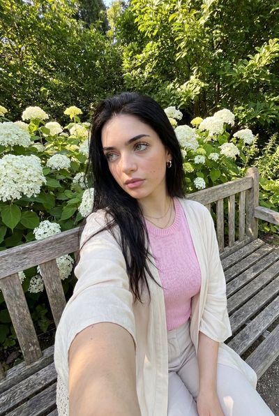 A woman takes a candid selfie on a wooden park bench surrounded by flowers