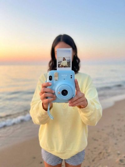 A person holds a baby blue Instax camera on a sandy beach at sunset.
