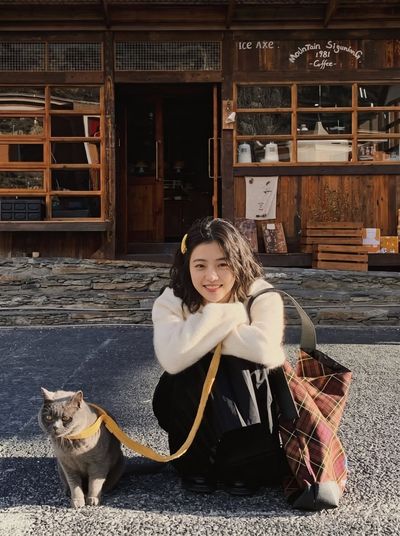 Young Woman Crouches With Gray Cat Outside Mountain Coffee Cabin