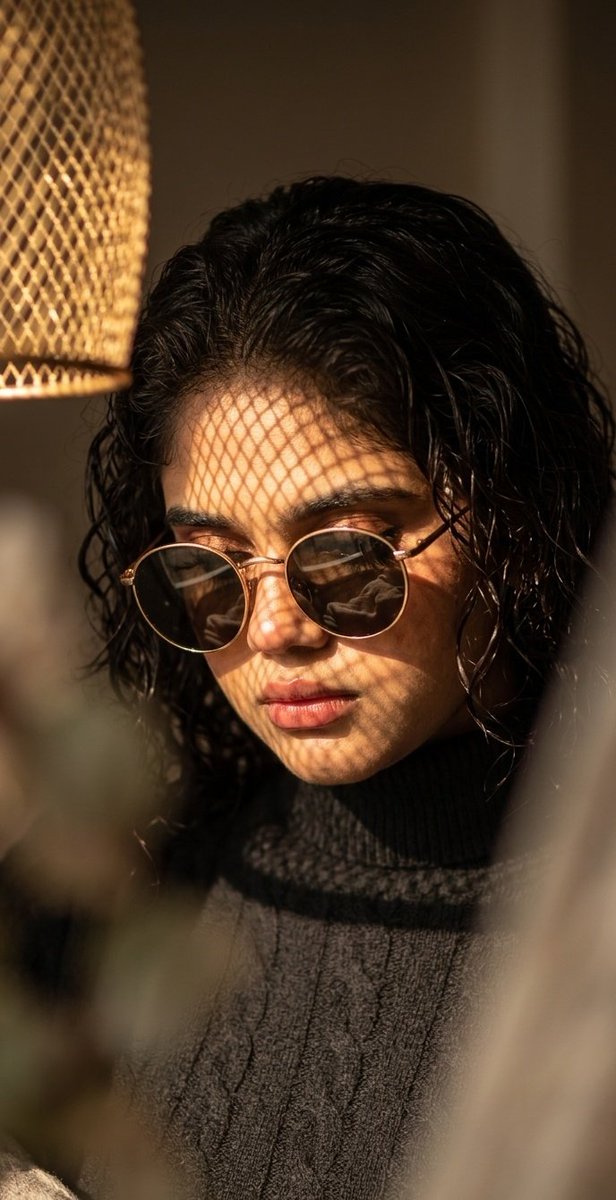A woman with gold sunglasses gazes downward in warm dappled sunlight.