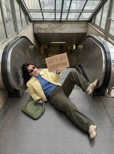 A young woman lies on an escalator holding a shopping sign in natural daylight.