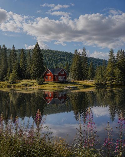 Red cabin beside a still alpine lake reflects clouds and mountains.