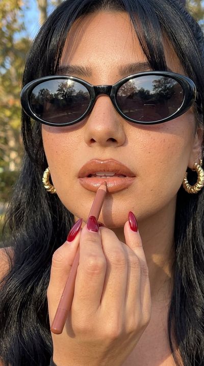 Woman applies caramel lip liner with gold hoop earrings in sunlight