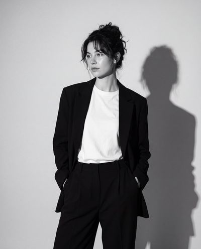 Woman in blazer stands with hands in pockets, shadow behind.