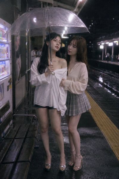 Two women share a quiet moment under a rainlit umbrella on a station platform