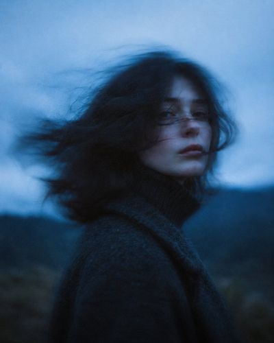 A woman in a dark wool coat turns amid windy twilight haze