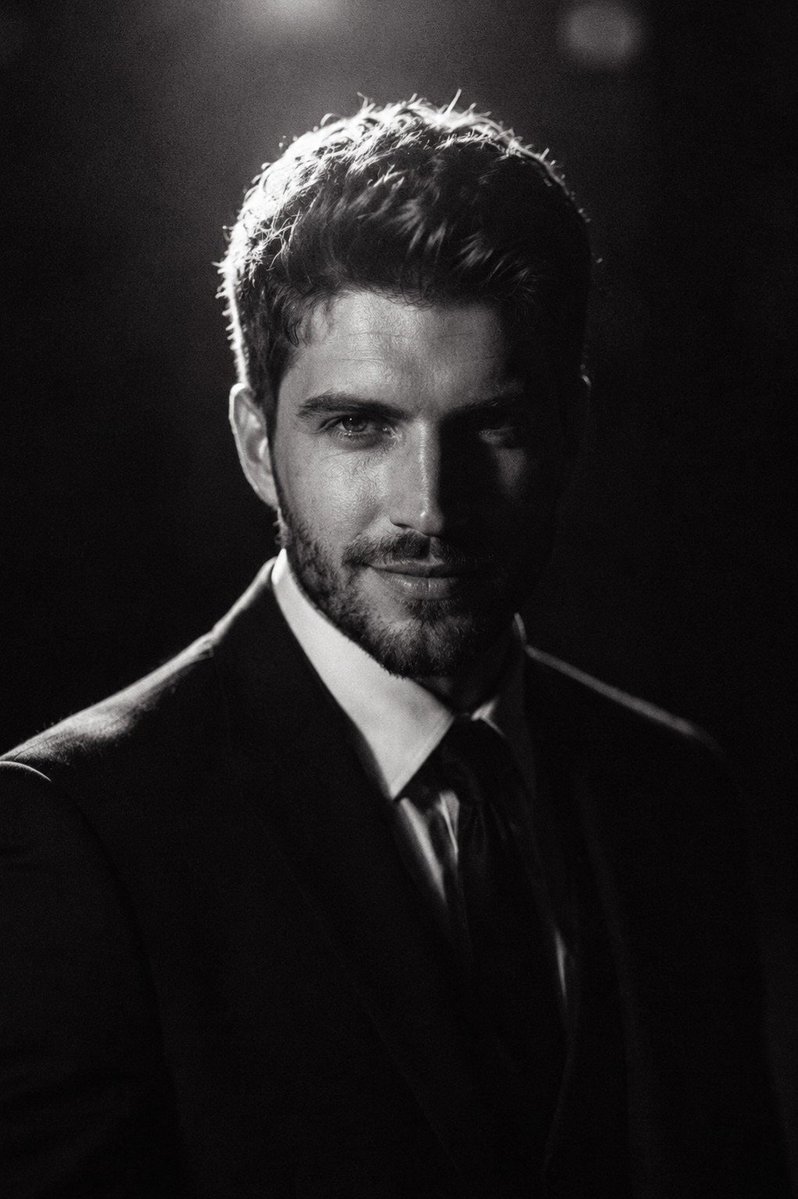 A young man in a black suit glows under dramatic low-key lighting.