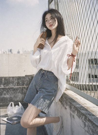 A woman in white leans by a fence with a glass bottle