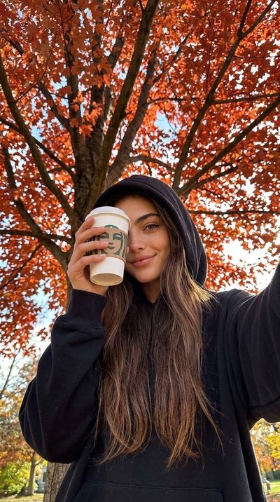Girl in black hoodie holds coffee under autumn tree