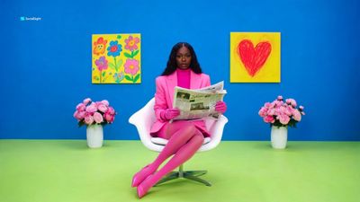 A dark-skinned model in pink reads a newspaper in a cobalt room.