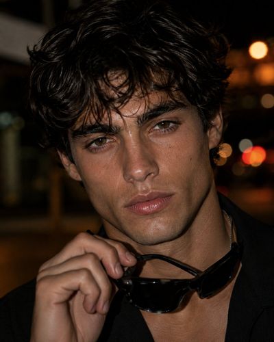 Young man with tousled hair holds sunglasses under warm city bokeh