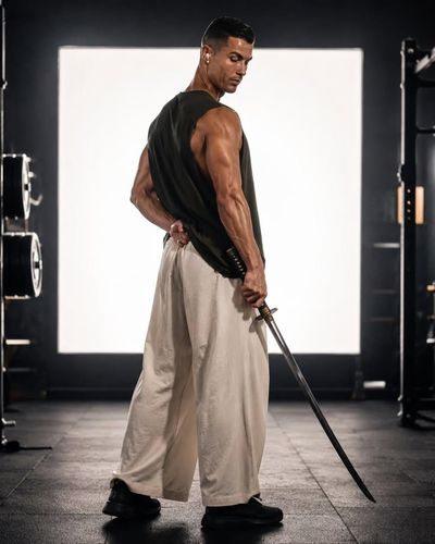 A muscular man in a gym turns his head while holding a katana