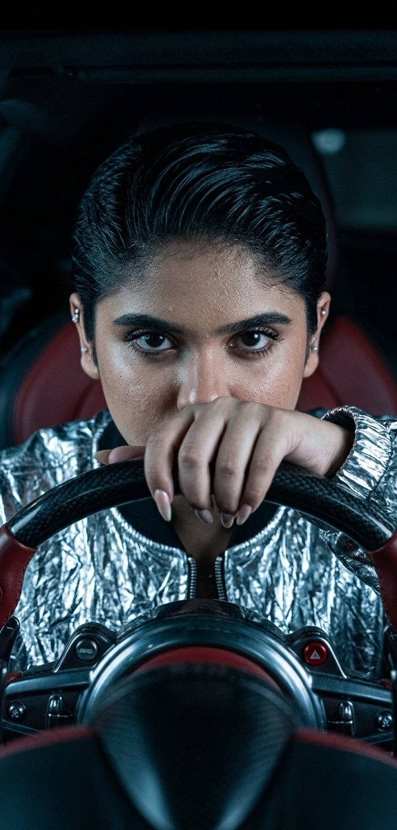 A woman with intense eyes grips the steering wheel inside a dark hypercar.
