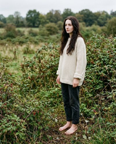 Slender woman in white sweater stands barefoot amid brambles