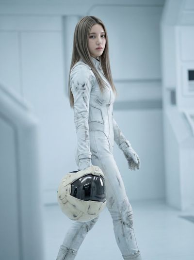 A young woman in a worn white pilot suit walks with a helmet