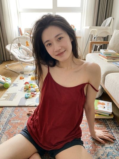 Woman in red top leans back in sunlit living room with baby items