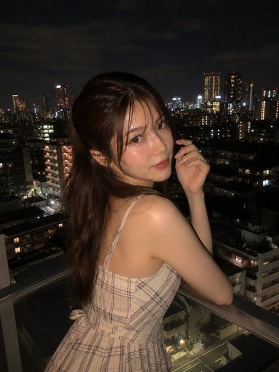 Young woman in cream plaid dress glances back from balcony at night