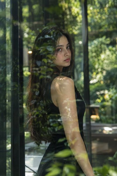 Woman in black dress gazes through reflective glass amid lush greenery