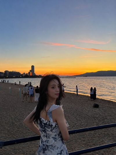 A woman in a blue floral dress looks back at a sunset beach.