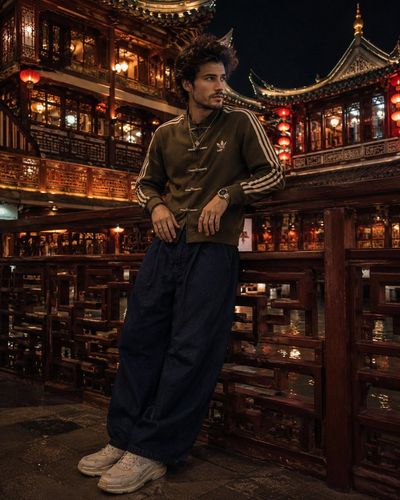 A man in olive Adidas jacket leans on Yu Garden balcony rails at night.