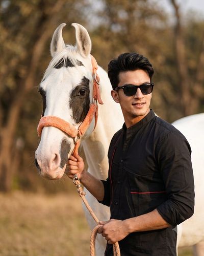 A man in black sunglasses gently leads a white horse in warm daylight