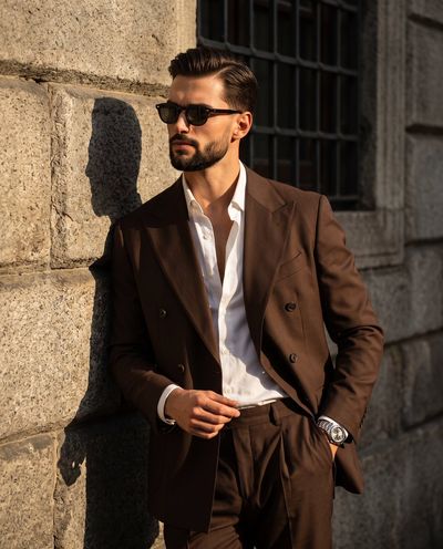 Confident man in chocolate suit leans against sunlit stone wall