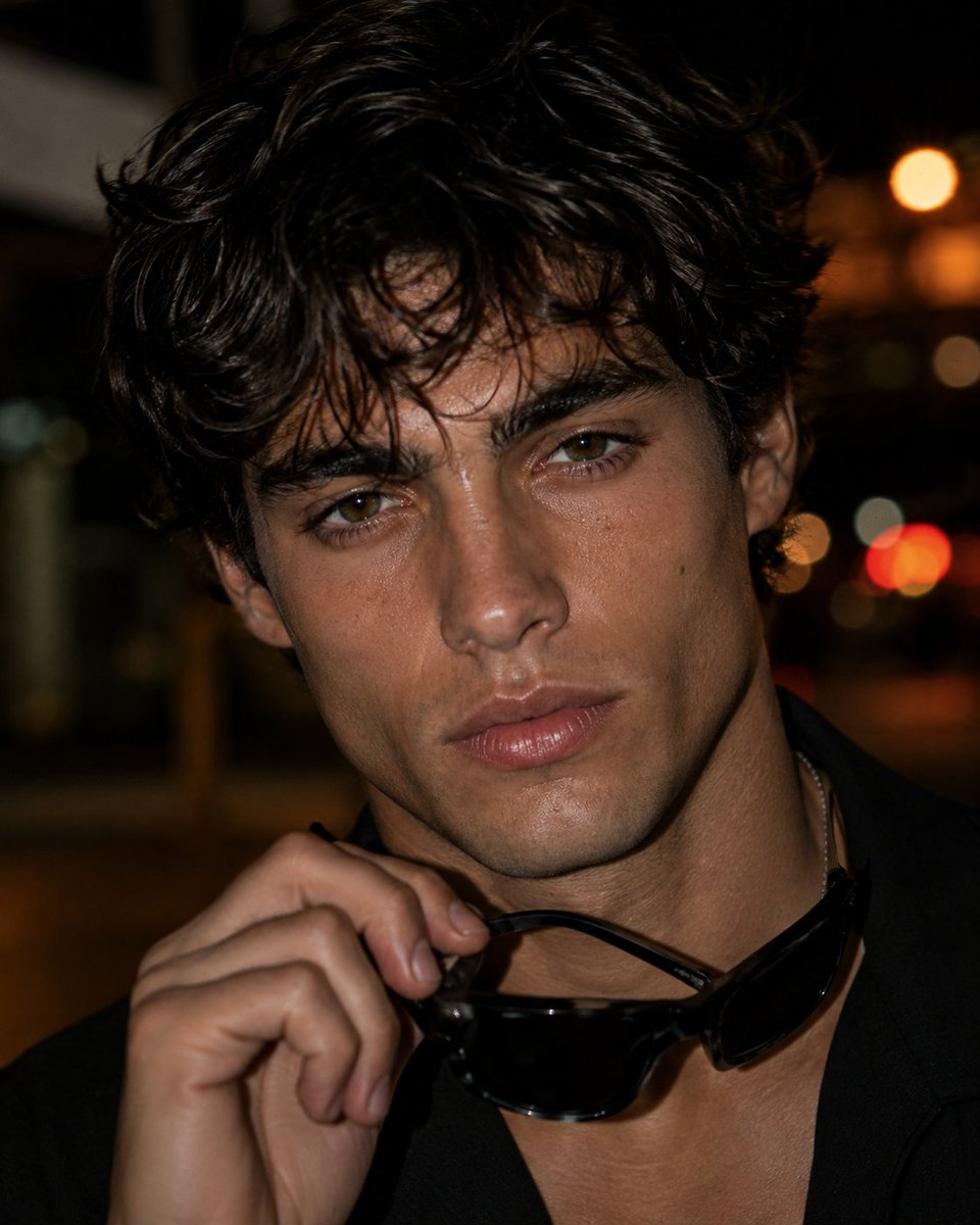 Young man with tousled hair holds sunglasses under warm city bokeh