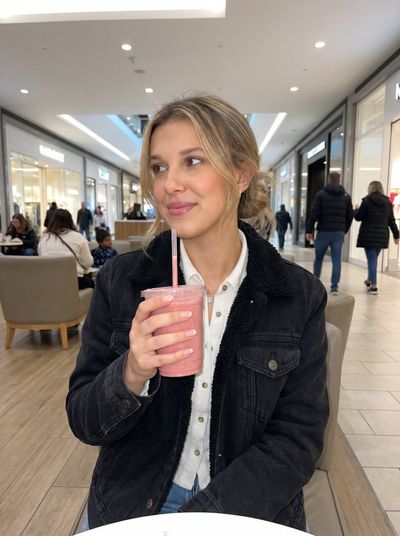 A young woman in a denim jacket smiles while holding a pink smoothie.