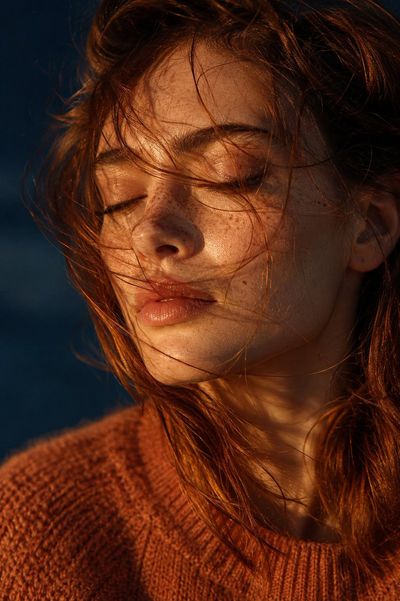 Wind-tossed auburn hair veils her serene freckled face in golden light