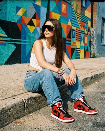 Young woman in baggy jeans and Air Jordans sits on graffiti-lined curb