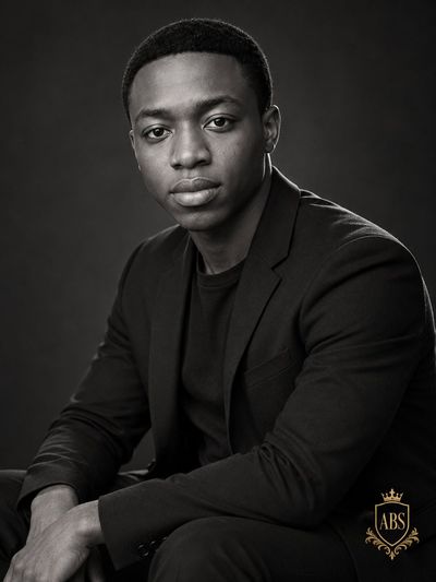 Monochromatic Elegance: Seated Man in Black Blazer with Gold Monogram