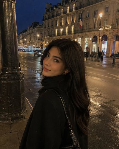 A woman in a black coat smiles under warm streetlights in Paris at night.