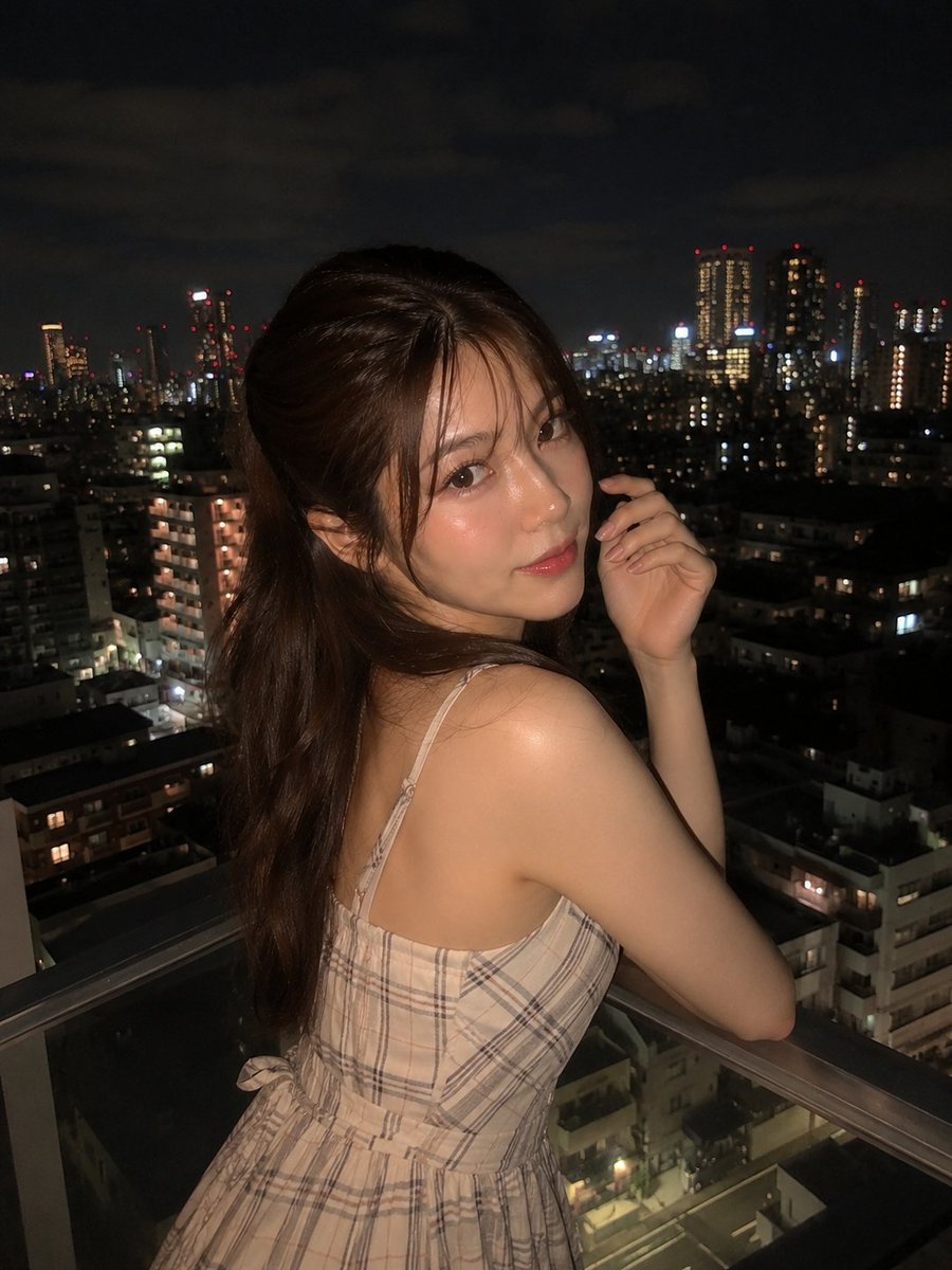 Young woman in cream plaid dress glances back from balcony at night