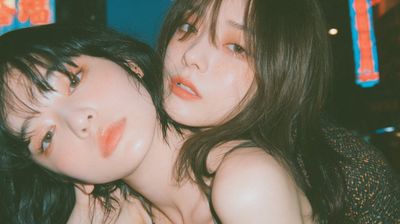 Two women crouch in neon-lit Hong Kong alley under azure sky