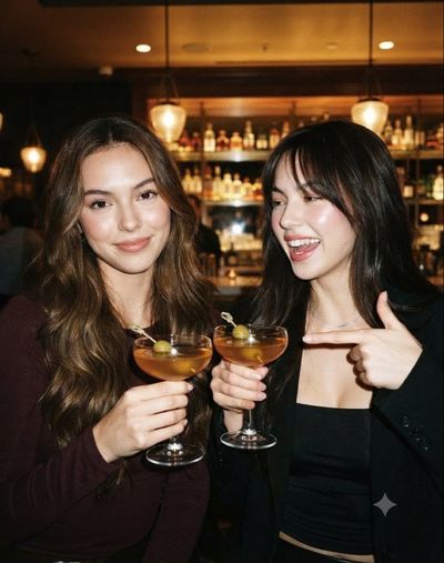 Two friends laugh over amber cocktails in a warmly lit upscale bar at night.