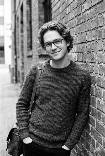 Young man with glasses smiles warmly in a brick alley