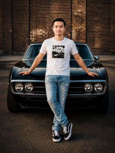 Young man leans against a glossy black muscle car in an urban lot