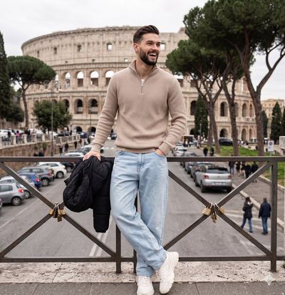 A smiling man in beige sweater leans against a railing near the Colosseum