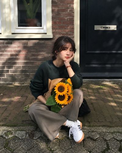 East Asian woman sits on a quiet sidewalk holding sunflowers